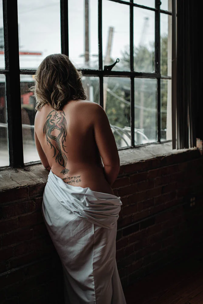 Woman with large back tattoo wrapped in a white sheet, leaning in the window, exposed brick.