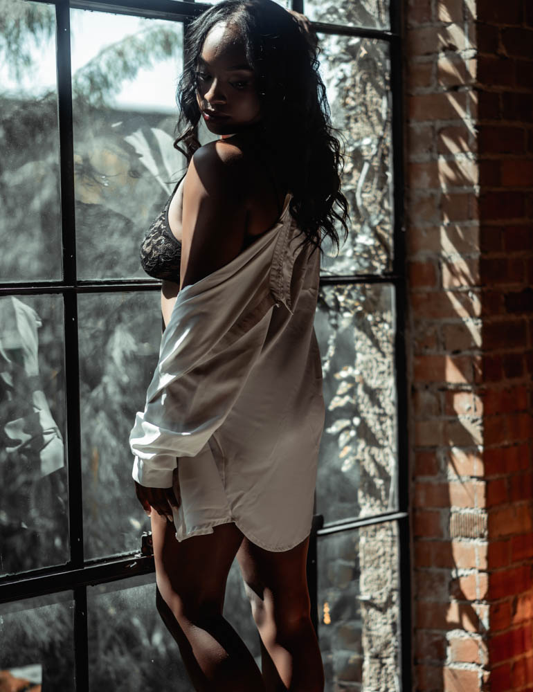 Woman standing in loft window with exposed brick on the walls, wearing a white button-up shirt and black lingerie