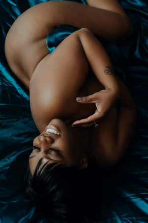 Black woman smiling, laying on turquoise silk sheets.