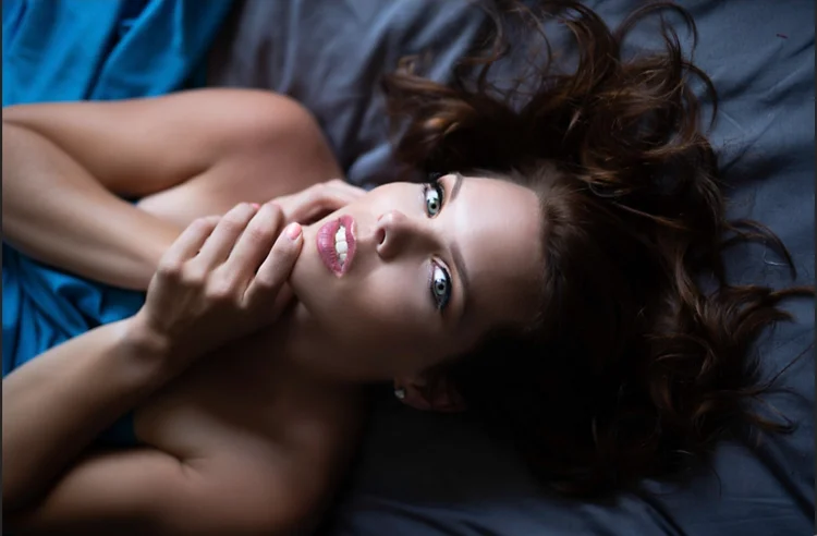 Woman laying on the bed, face up with red hair, hands on the cheek, looking into the camera, wrapped in a blue sheet