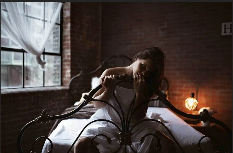 Woman sitting on antique bed wrapped in a sheet near a window with exposed brick.