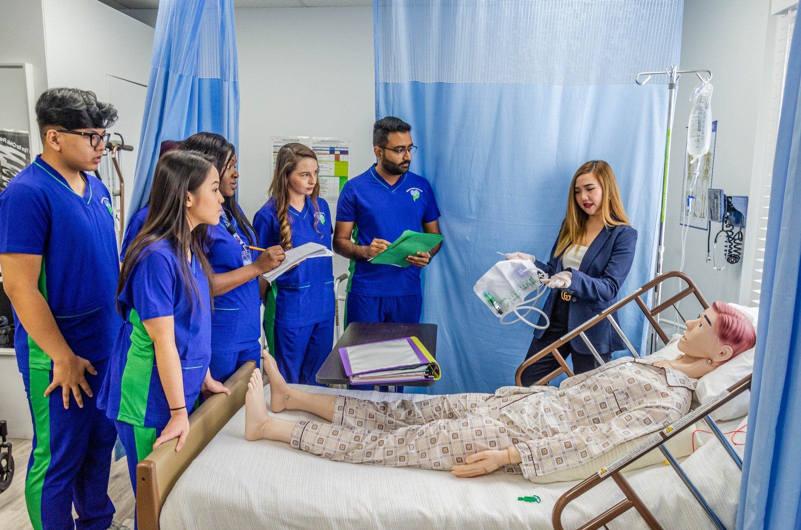 AHCS students taking notes as an instructor demonstrates a catheter bag on a patient simulation manikin in the practice lab