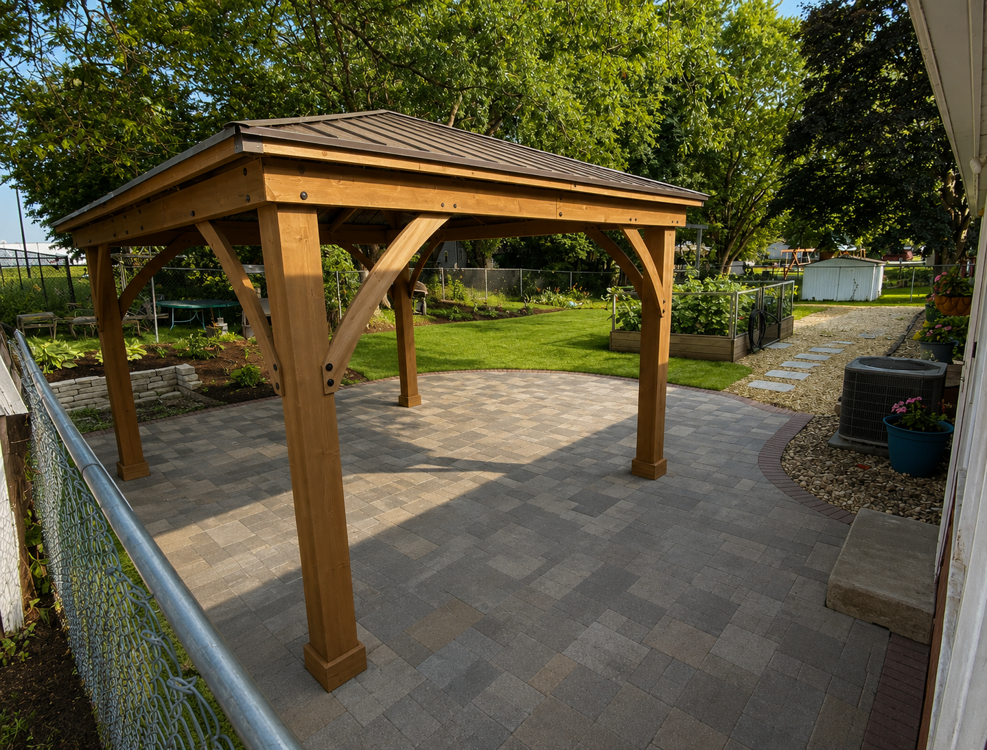 Open timber-frame pavilion with a shingled hip roof on a paver patio beside a railing, with lawn and trees behind.