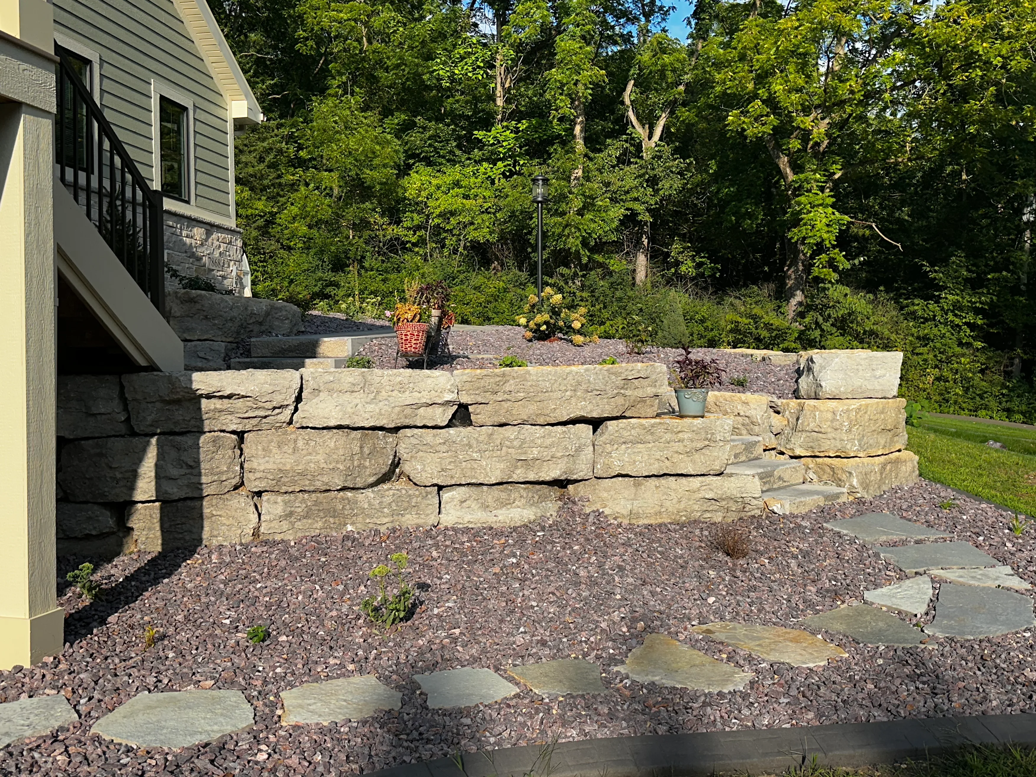 Two-tier stone retaining wall creating planted terraces on a wooded slope, with stepping stones through rock mulch below.