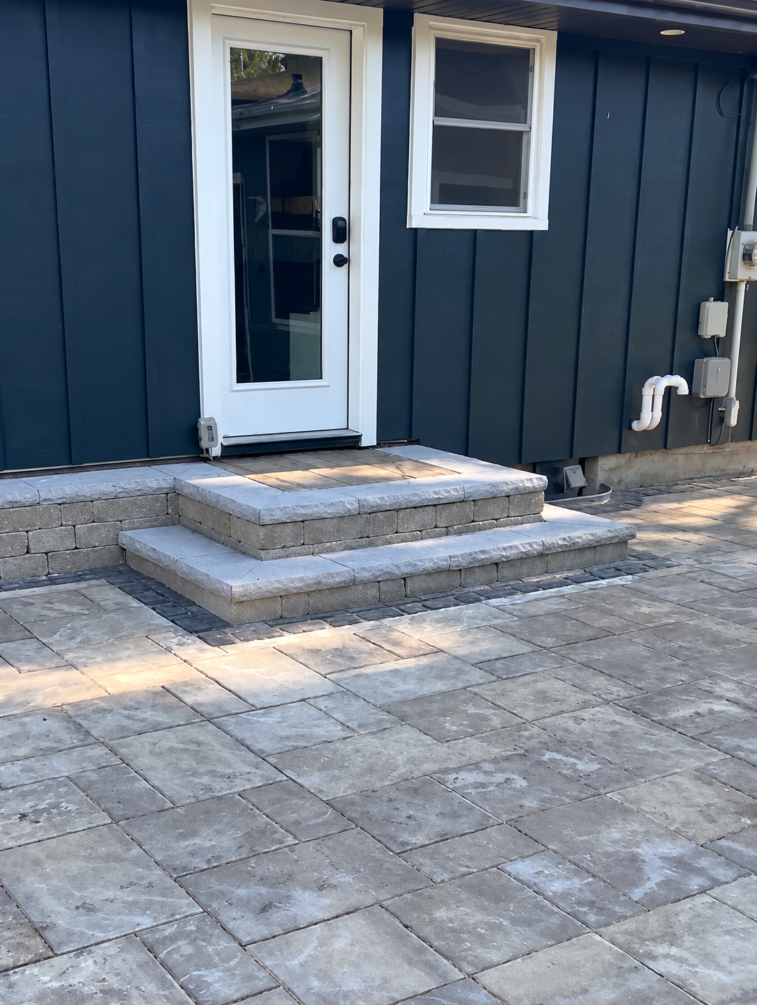 Round paver patio with a stone fire pit and white Adirondack chairs in a fenced back lawn.
