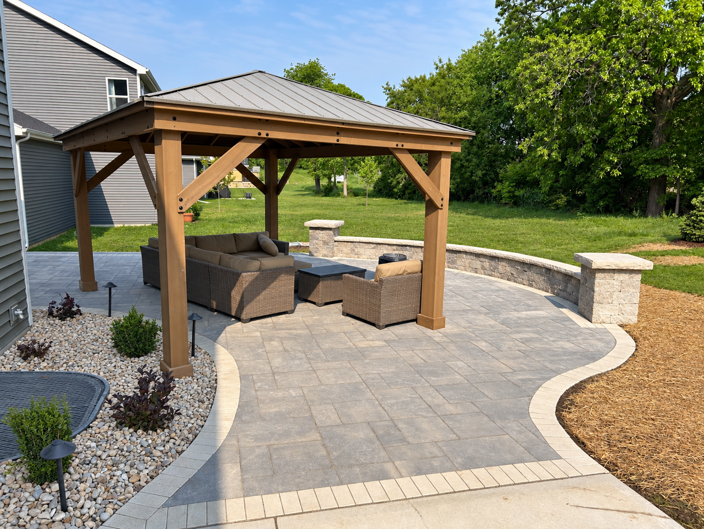Cedar-toned timber pavilion with a hipped metal roof sheltering outdoor furniture on a paver patio, with a curved block seat wall and lawn beyond.