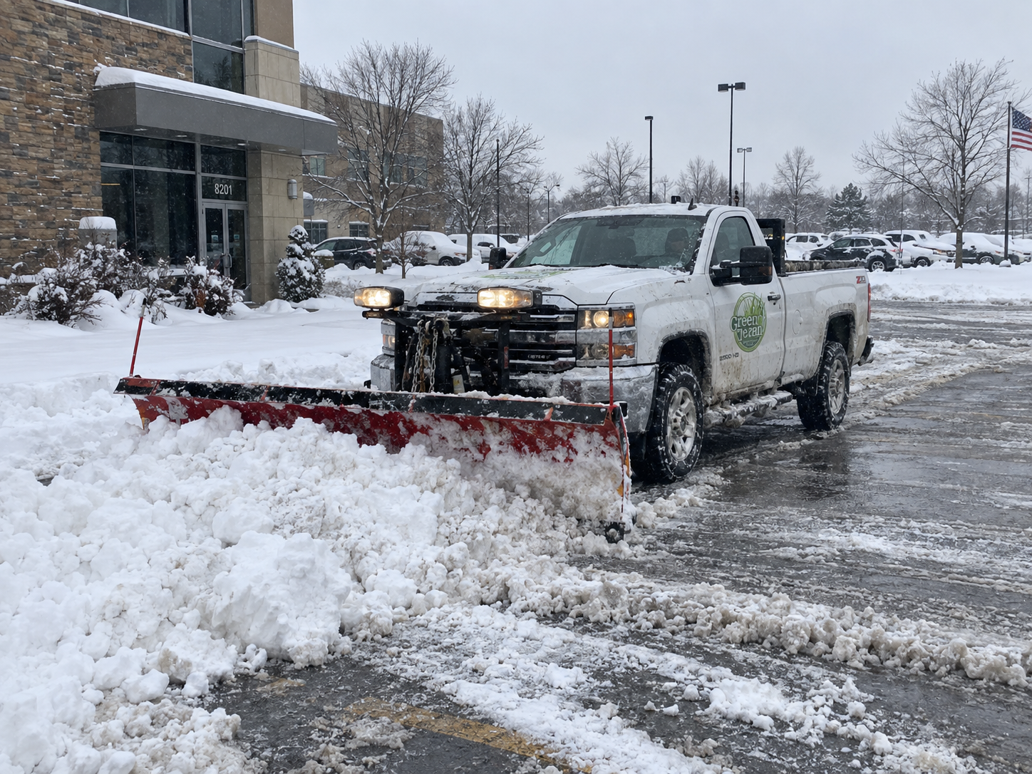 White pickup with a red plow pushing snow across a commercial parking lot during active snowfall, with a building and light poles behind.