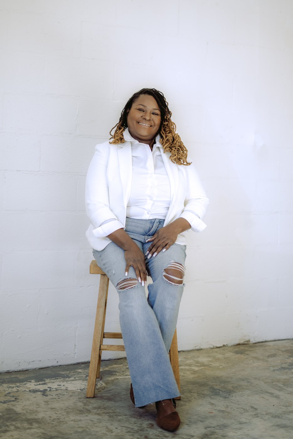 Starr Clinton standing against a white wall in a white blazer and jeans