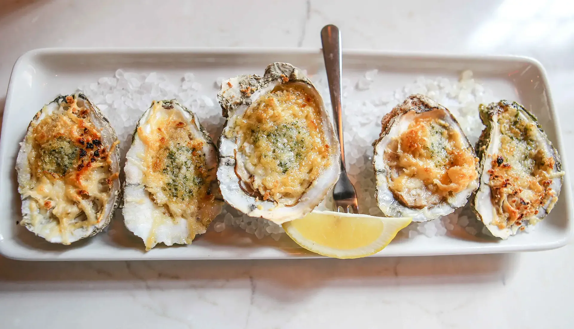 Baked oysters topped with garlic herb butter, served on a bed of rock salt with a lemon wedge.