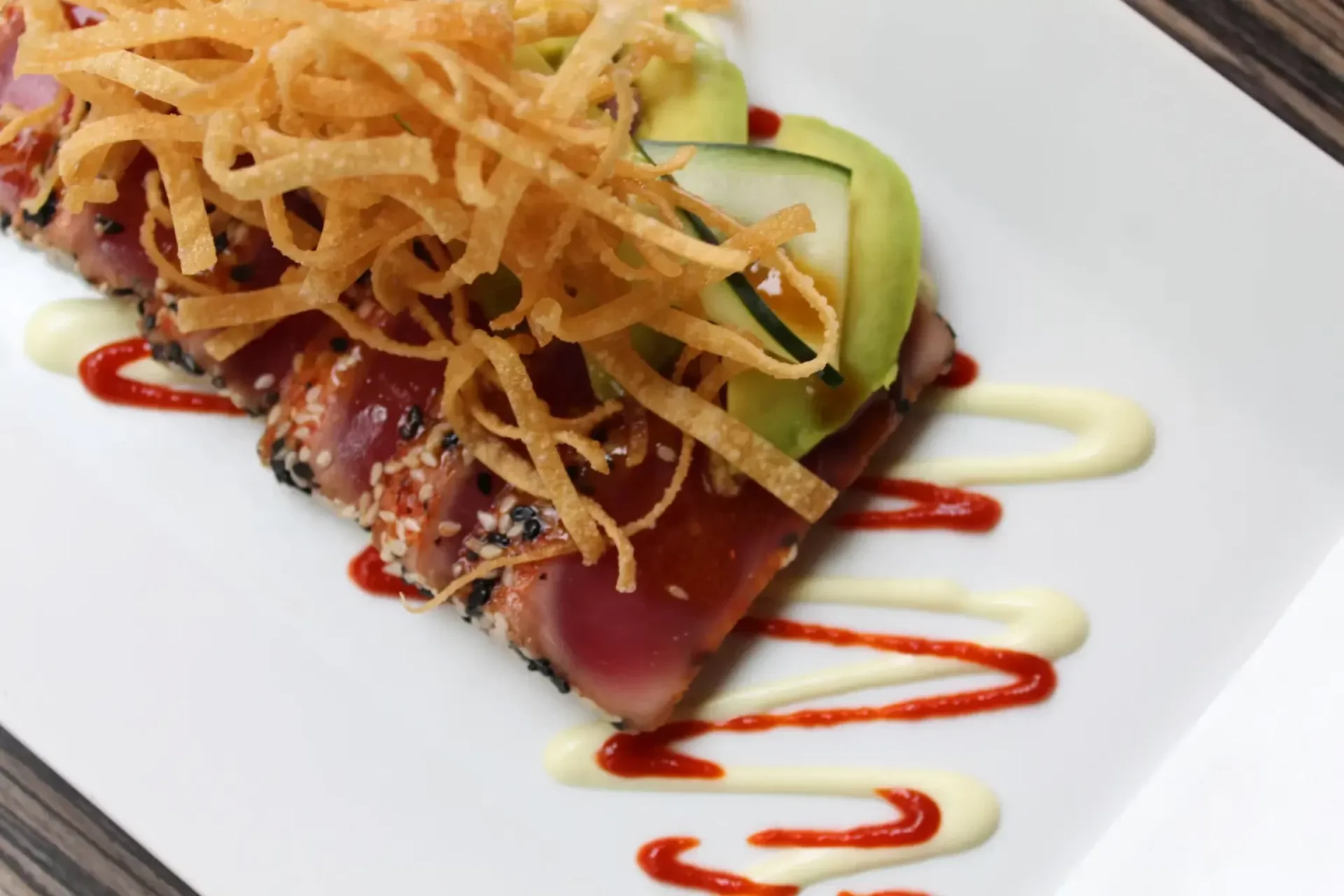 Ahi tuna topped with crispy fried onions and avocado slices, drizzled with spicy and creamy sauces on a white plate.