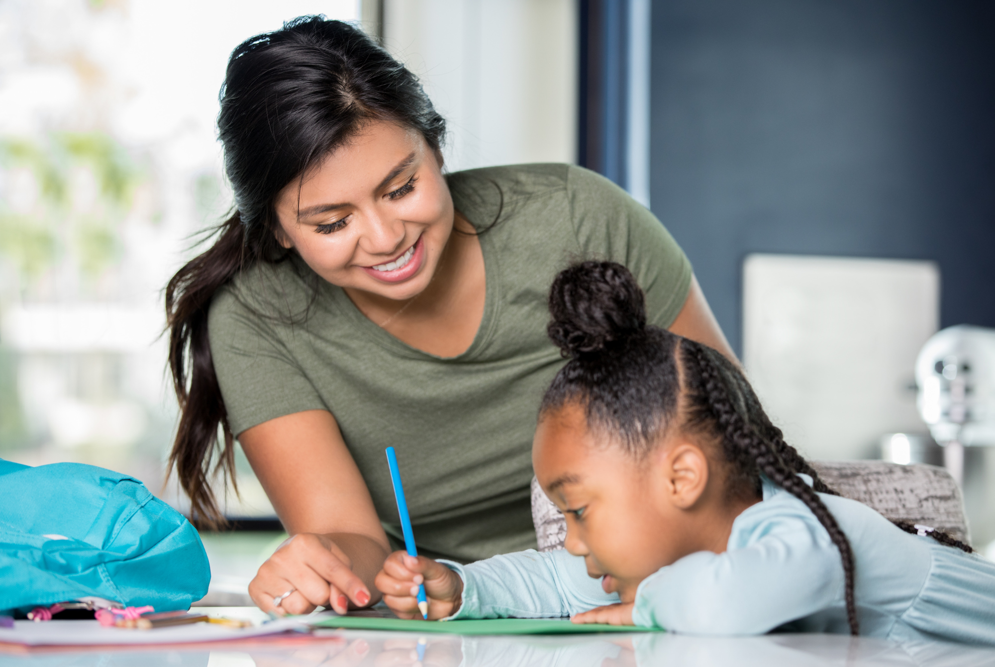 A teen or tween girl with olive skin and dark hair wearing a green shirt colors with a young girl with olive skin and black hair while babysitting.
