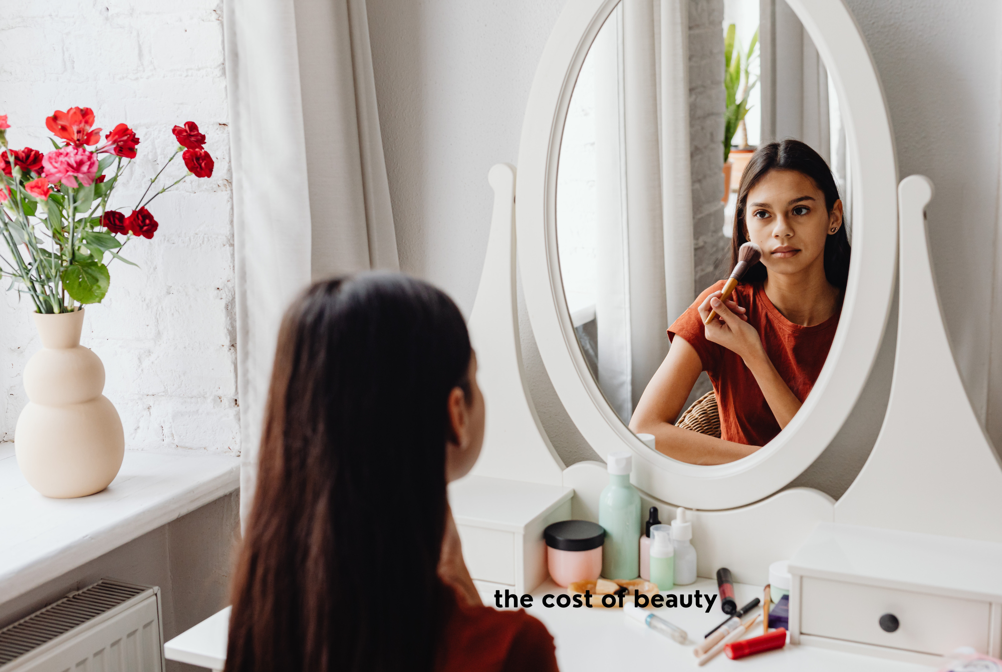 A tween or teen girl with dark hair and skin sits in front of a white oval mirror spending too much time, energy, and money getting ready for the day.