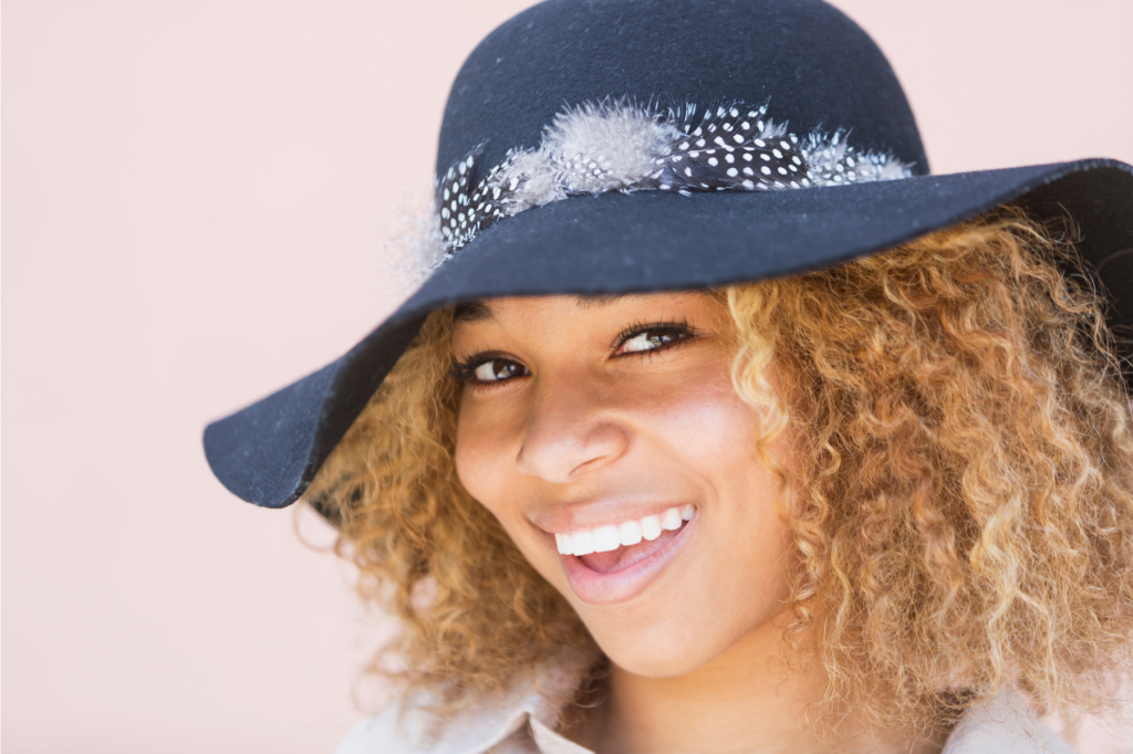 A black teen or tween girl with light brown hair smiles brightly in a black wide brimmed hat.