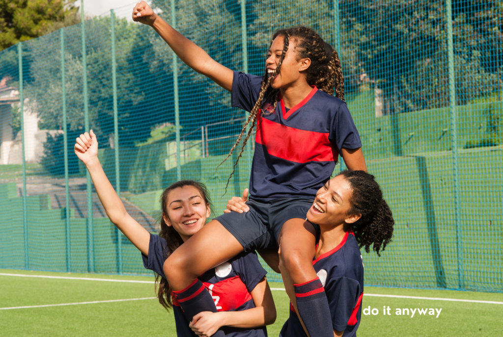 Two teen or tween soccer player girls hold a teammate on their shoulders.