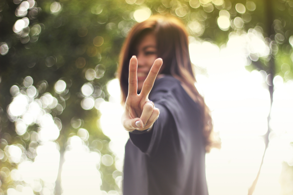 A tween or teen girl with brown hair and skin wearing a gray shirt stands outside near trees  holding up a peace sign to the camera.