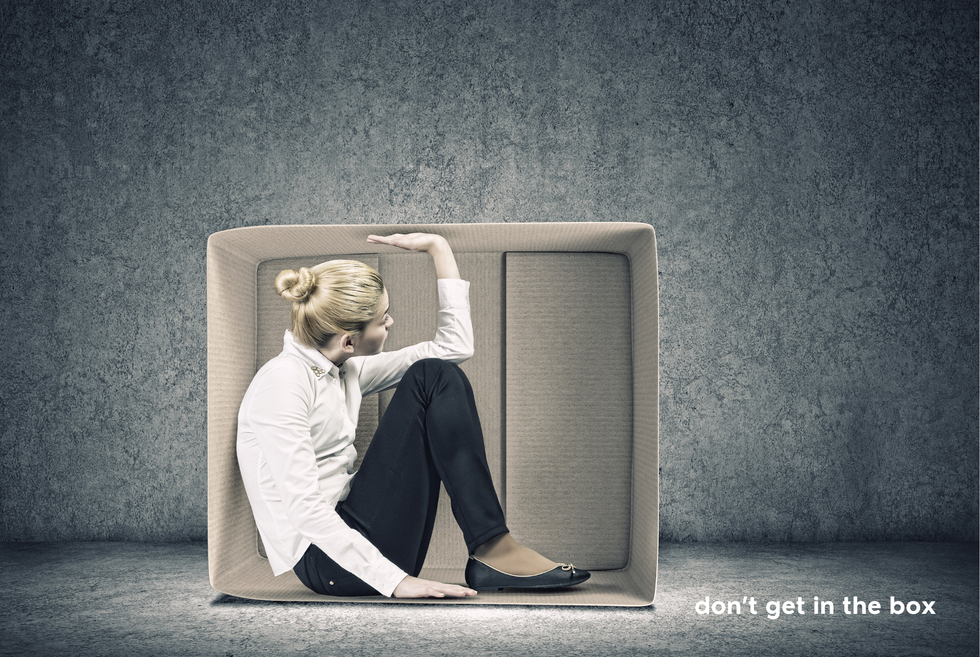 A teen or tween girl sits squished inside a box sitting on its side, pushing against the top.