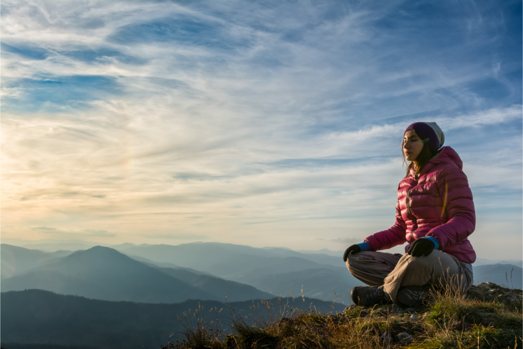 A tween or teen girl with brown hair and tan skin wearing a beanie and pink coat and tan pants sits cross legged on top of a cliff overlooking a mountain range in the daylight.