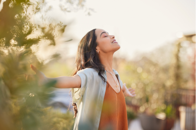 A tween or teen girl with tan skin and brown hair stands outside with her head back, eyes closed, and arms out wide as she copes with change.