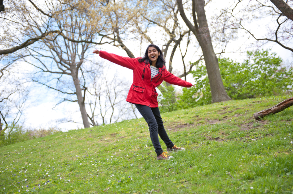 A tween or teen girl with dark hair and skin stands on a grassy hill wearing a red coat and jeans and extending her arms wide in gratitude.