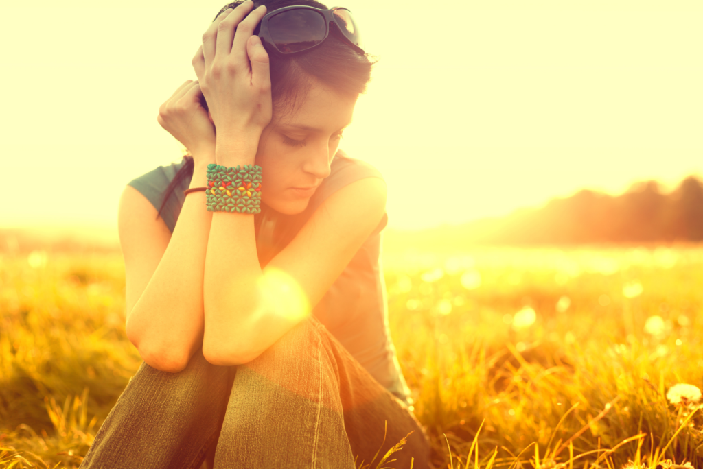 A yellow filtered photo of a tween or teen girl sitting in the grass, resting her head on her hands, looking stressed about things she can't control.