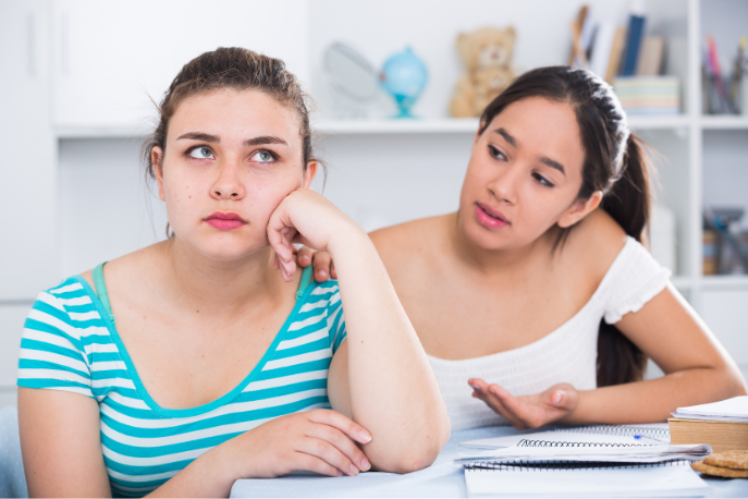 A tween or teen girl with dark hair wearing a white shirt apologizes to another tween or teen girl with brown hair wearing a teal striped shirt.