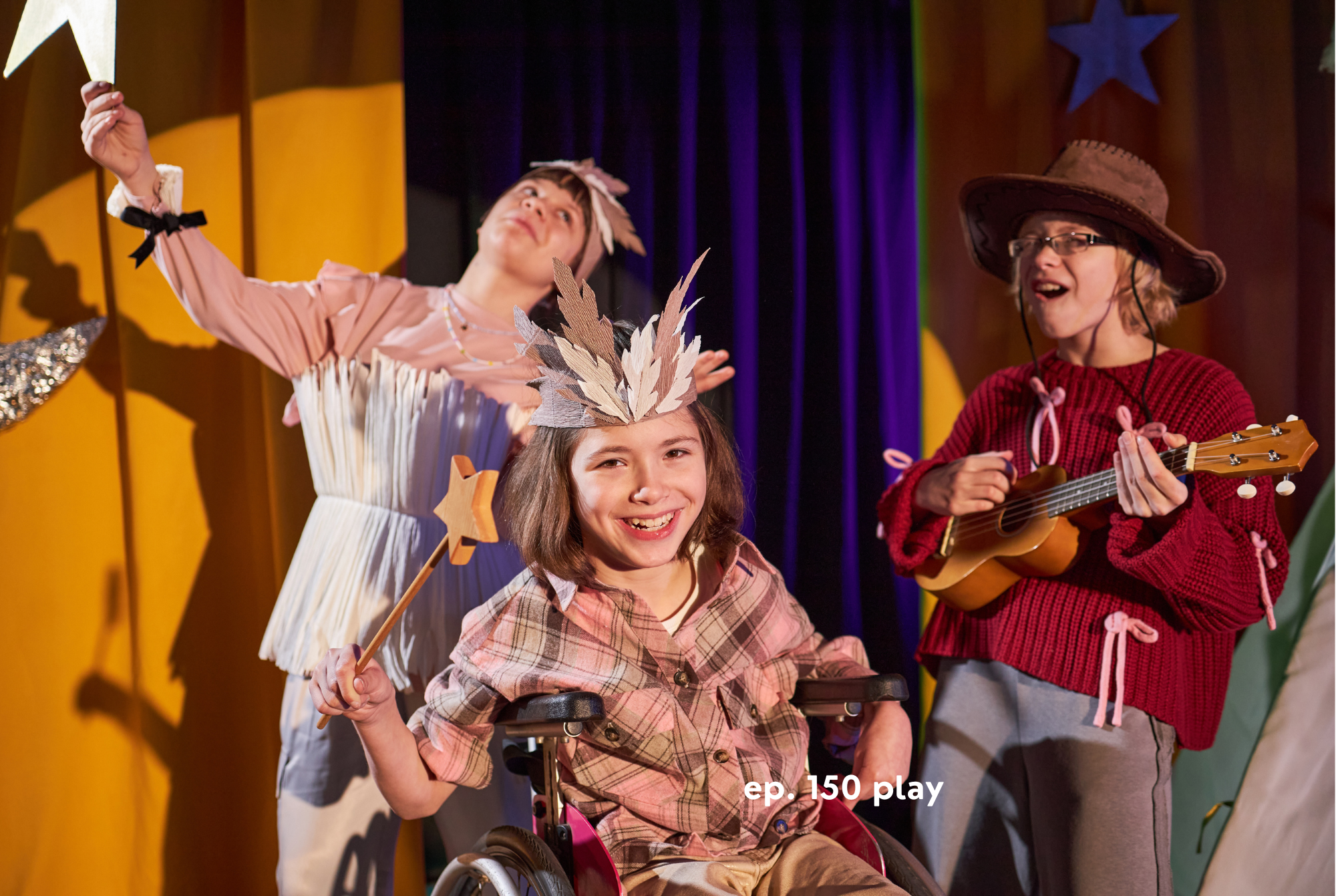 A tween or teen girl who uses a wheel chair, and two girls standing behind her, all wearing whimsical costumes, act out a scene together