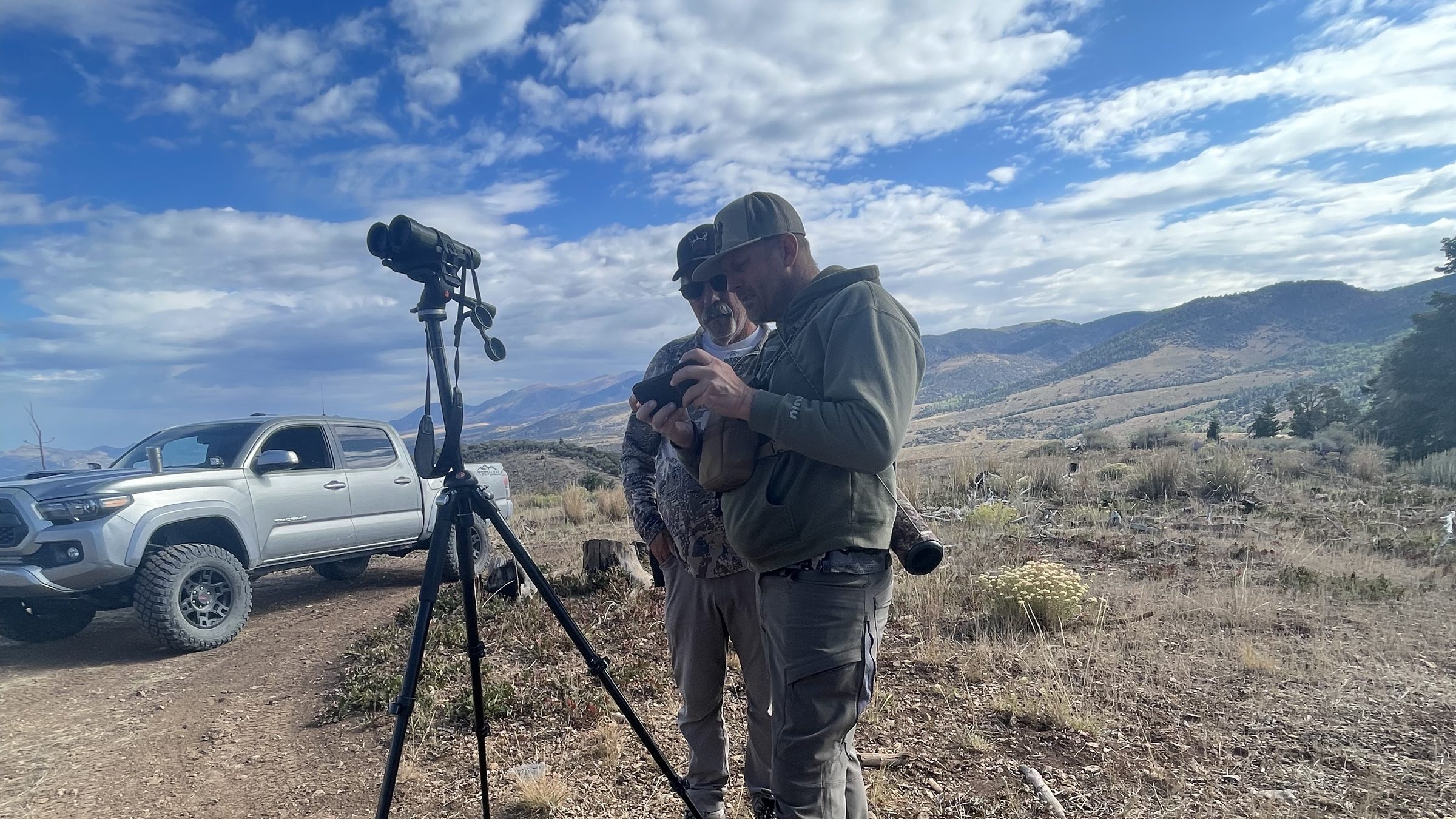 Hunters glassing with a spotting scope on a tripod from a ridge under a wide sky