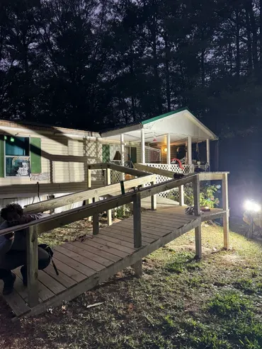 A person crouches on a wooden access ramp extending to a manufactured home with a brightly lit porch against a dark treeline.