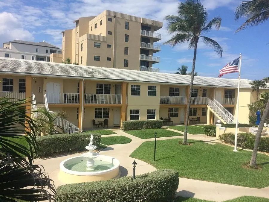 A two-story yellow building with balconies, stairs, a courtyard fountain, green grass, palm trees, and an American flag.