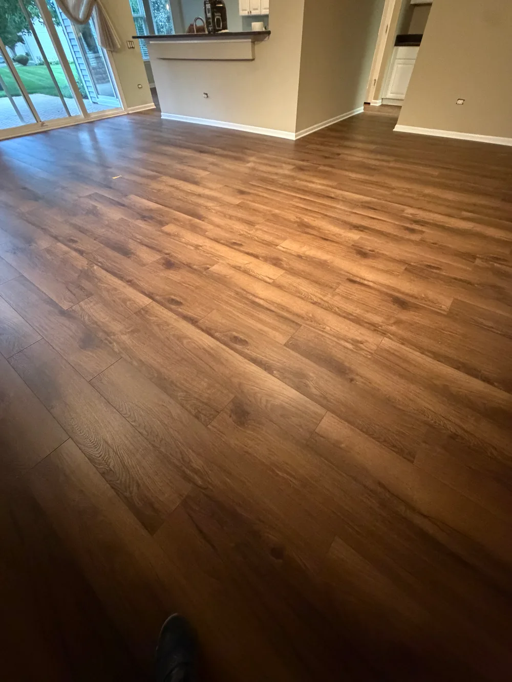 Newly installed medium-brown wood-look laminate flooring fills a bright living space, extending towards a patio and kitchen.
