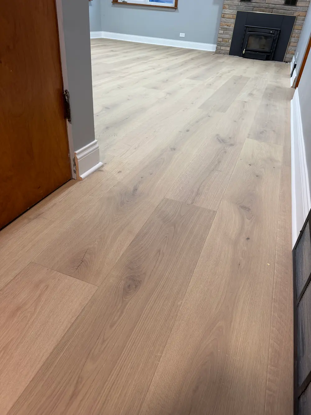 Light wood-look laminate flooring in a room with gray walls, white baseboards, a brown door, and a brick fireplace.
