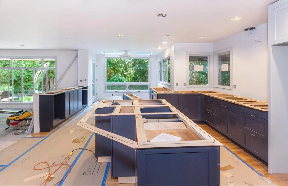 Spacious kitchen mid-renovation showing dark blue base cabinets, a large island outline, and natural light from many windows.