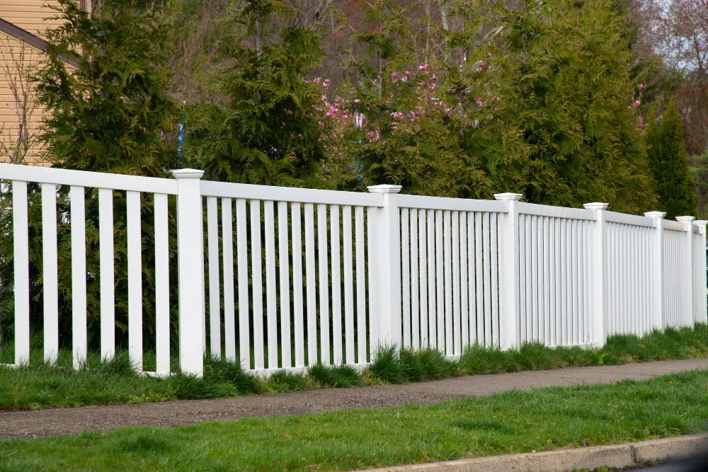 Modern dark gray louvered fence and matching gate extends along a paved sidewalk and gravel driveway under a blue sky.