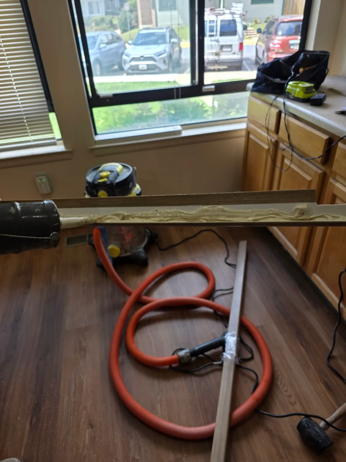 Glue being applied to trim on a wood floor, with a shop-vac, kitchen cabinets, and outdoor cars visible through a window.