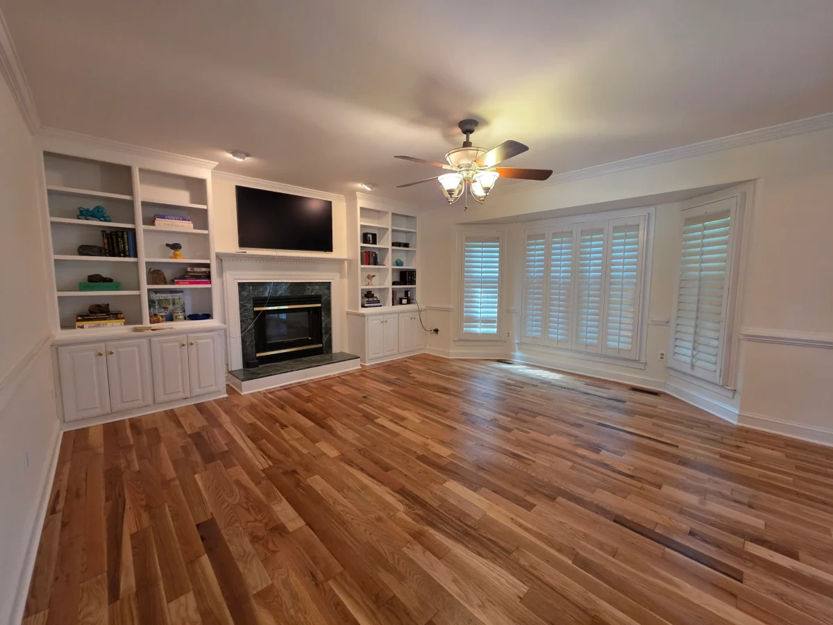 An empty living room with warm hardwood floors, white built-in bookshelves, a fireplace with TV, and bay windows with plantation shutters.