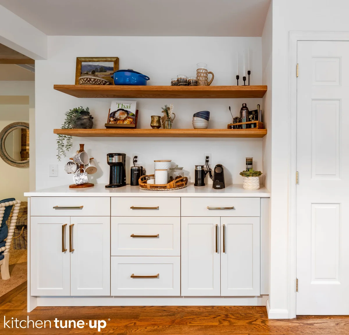 A stylish white kitchen features open wooden shelves adorned with decor, a coffee bar, and white base cabinets with bronze hardware.