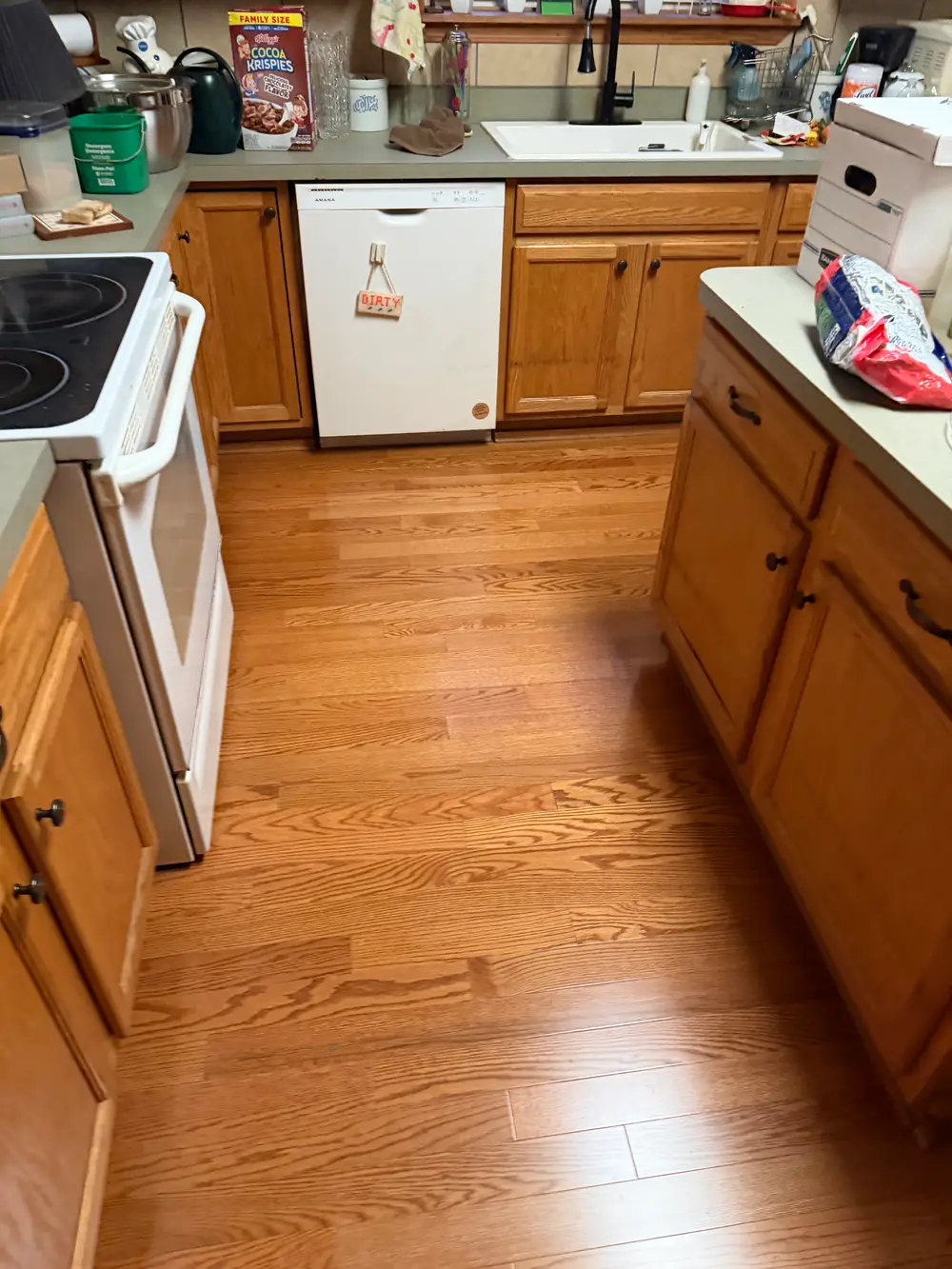 View of a home kitchen with honey-toned hardwood floors, oak cabinets, a white dishwasher, an electric range, and a sink.