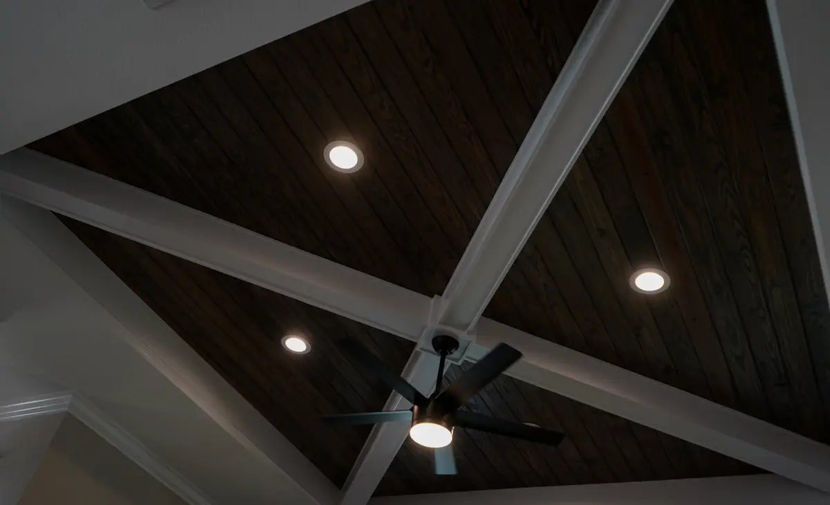 A beautiful dark wood plank ceiling with white geometric beams, multiple recessed lights, and a contemporary black ceiling fan.