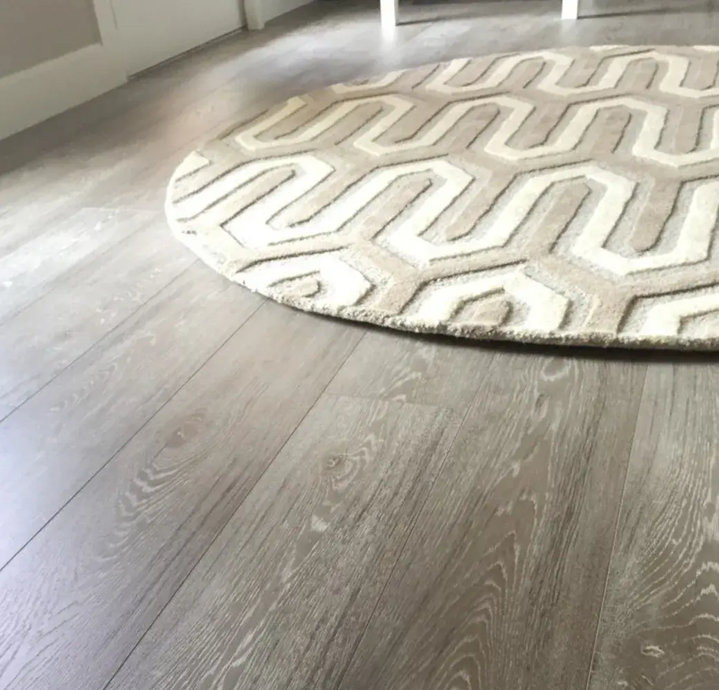Angled view of light grey wood plank flooring featuring a round cream and beige rug with a bold geometric pattern.