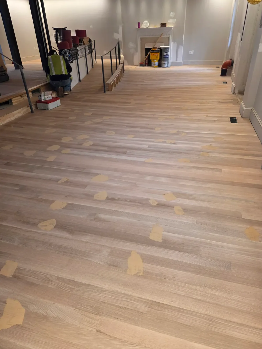 Light hardwood floor being prepped with beige filler patches, a worker, construction tools, and a fireplace.