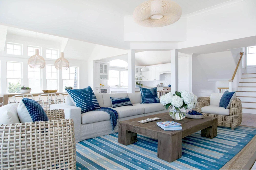 A serene coastal-themed living room with white sofas, blue accents, a rustic coffee table, striped rug, and wicker chairs.