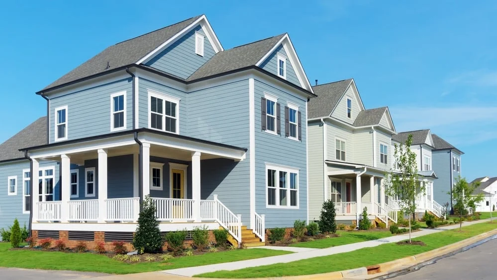 A neat row of newly built, pastel-colored suburban houses with inviting front porches, green lawns, and sidewalks under a clear sky.