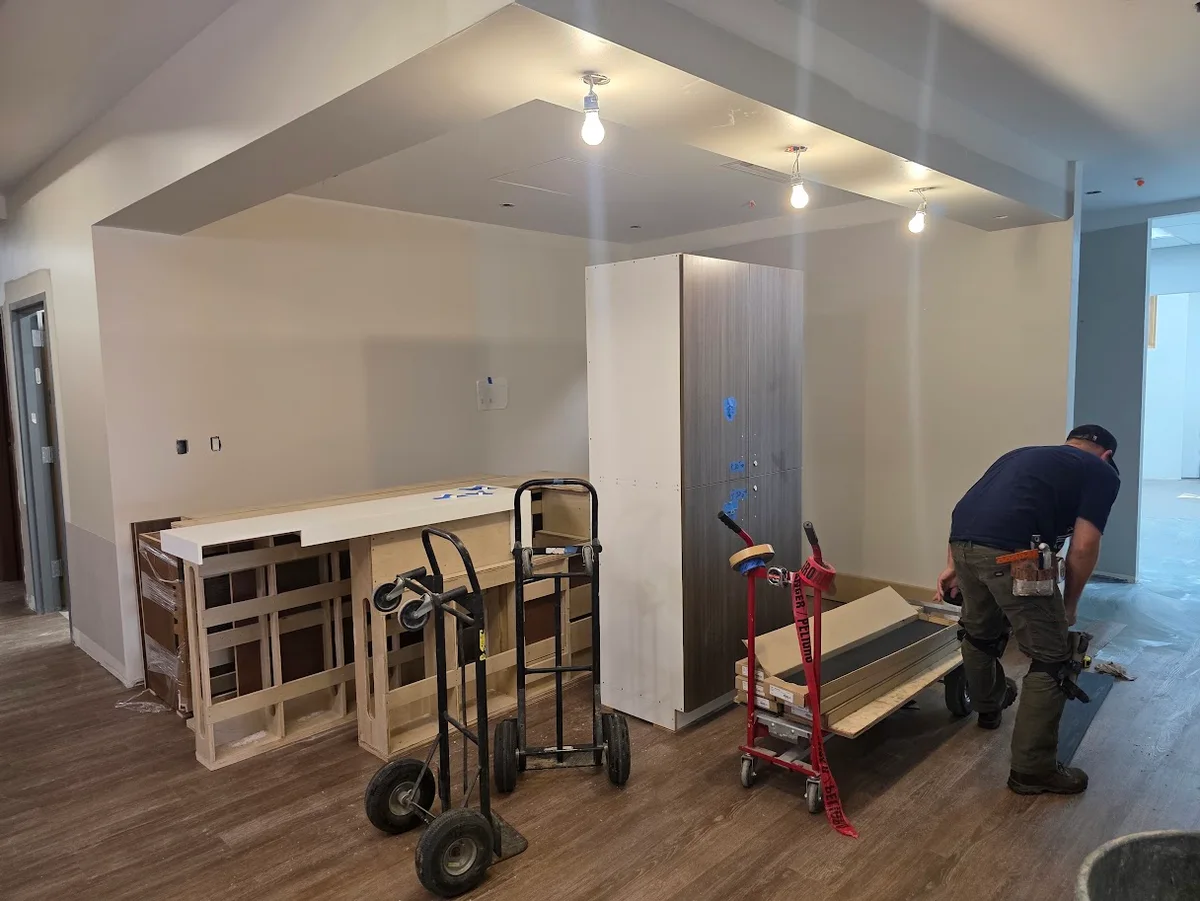 A construction worker installs flooring in a room with unfinished cabinets, boxes of materials, and utility carts.