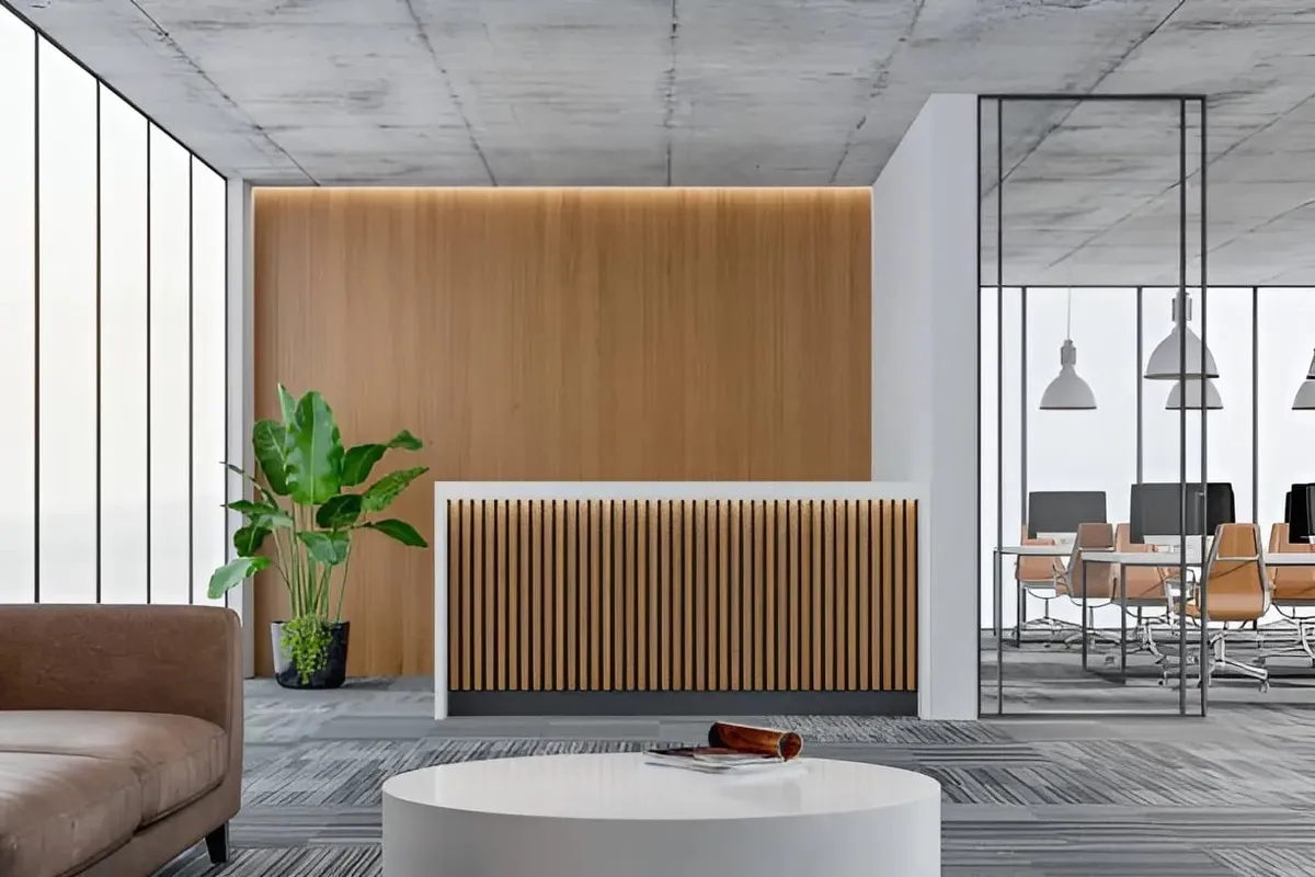 A modern office reception area features a wooden accent wall, a slatted reception desk, a lounge sofa, and a glass partition to an open workspace.