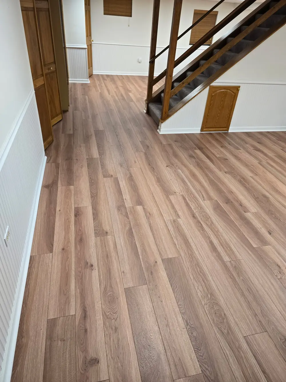 Newly installed light brown wood-look flooring extends through a room with white walls, wainscoting, and a wooden staircase.