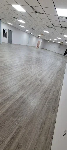 Spacious, empty room with light grey wood-look vinyl flooring, white walls, and a grid ceiling with bright lights.