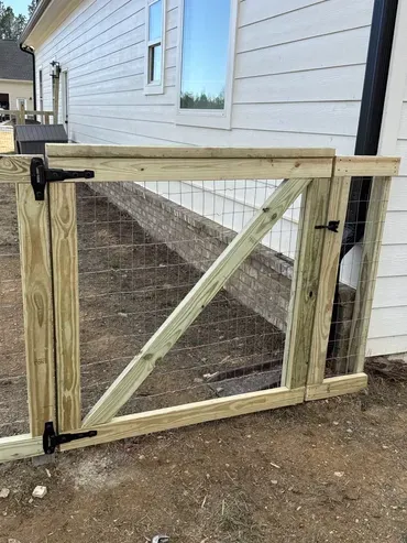 A newly constructed light wood gate with wire mesh, black hinges, and a latch, installed next to a white house with a brick wall.