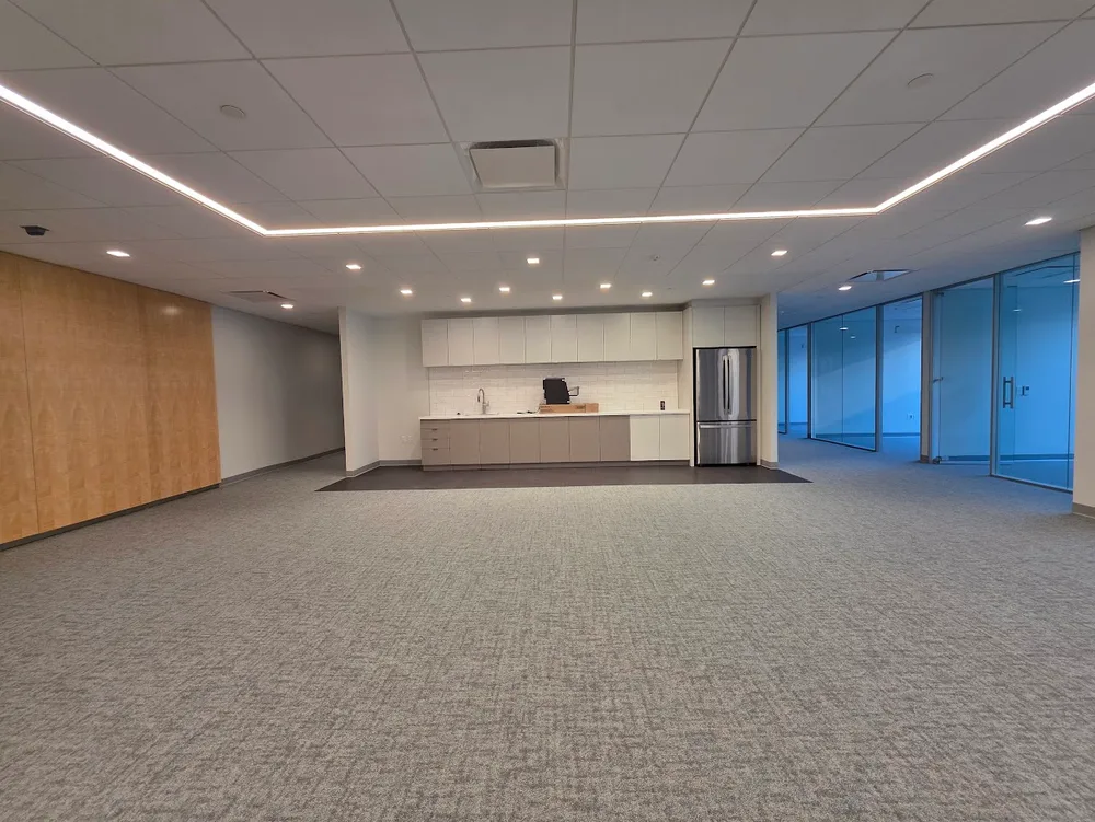 Empty modern office with a kitchenette, stainless steel fridge, gray carpet, wood wall, and glass-paneled office rooms.