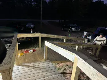 Workers apply finishing touches to a newly constructed wooden accessibility ramp and deck under artificial lights at night.