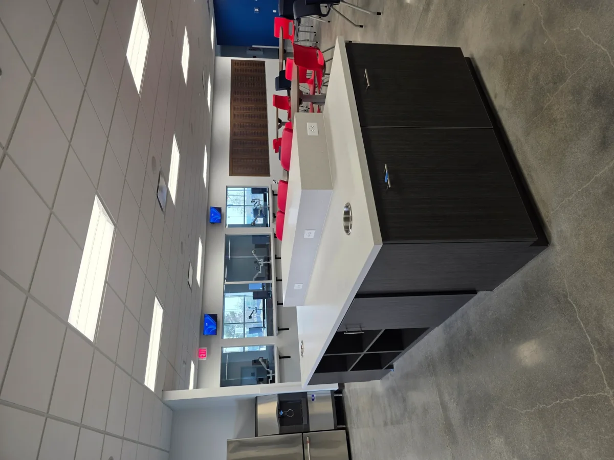 A modern office breakroom features a large kitchen island, concrete floors, red chairs, and glass-partitioned workspaces.